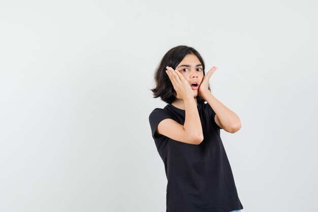 Menina de camiseta preta, segurando as palmas das mãos nas bochechas e olhando surpresa, vista frontal.