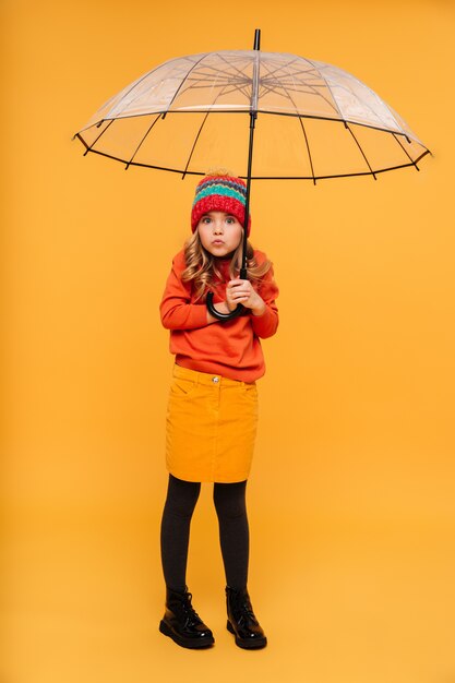 Menina de camisola e chapéu, escondendo-se atrás do guarda-chuva e olhando para a câmera sobre laranja