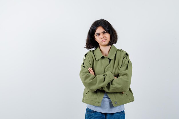 Menina de casaco, camiseta, jeans em pé com os braços cruzados e parecendo confiante, vista frontal.