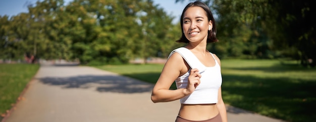 Foto grátis menina de fitness asiática sorridente segurando uma toalha no ombro exercitando-se no parque suando depois do treinamento