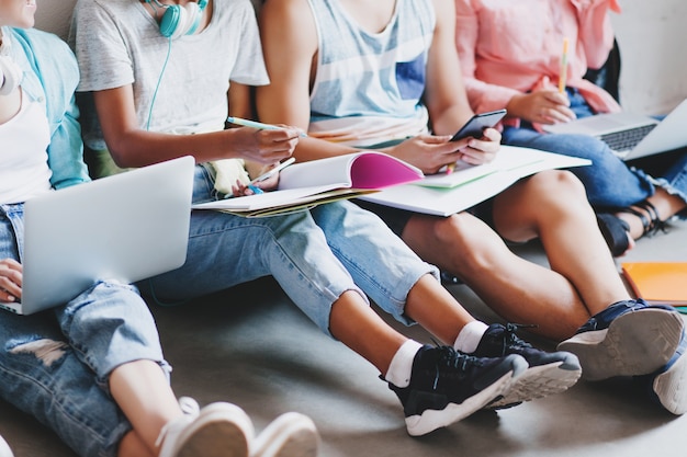 Menina de jeans e tênis preto, escrevendo uma palestra em um grande livro, sentada no chão com amigos da faculdade. Jovem digitando mensagem no telefone, enquanto outros alunos trabalham com laptops.