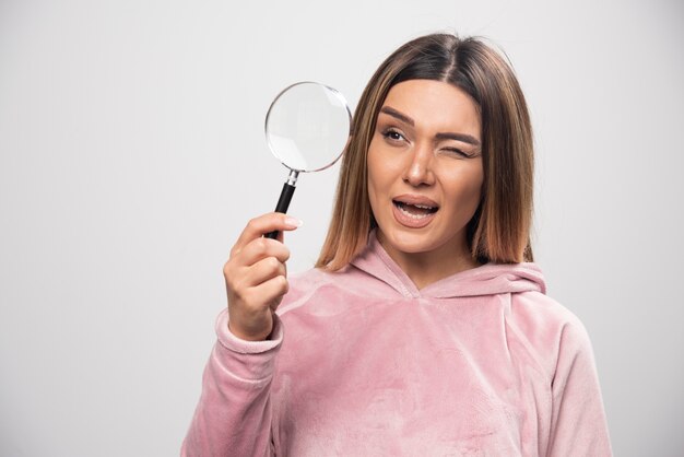 Menina de moletom rosa colocando uma lupa no olho e olhando através dela.