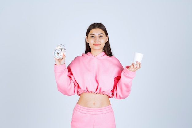 Menina de pijama rosa segurando um despertador e uma xícara de café.