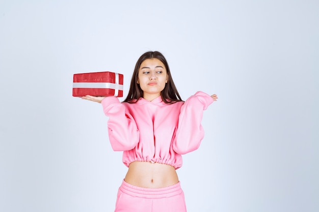 Menina de pijama rosa segurando uma caixa de presente retangular vermelha e parece indiferente.