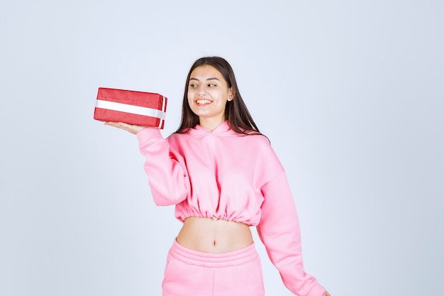 Menina de pijama rosa segurando uma caixa de presente retangular vermelha e parece satisfeita.