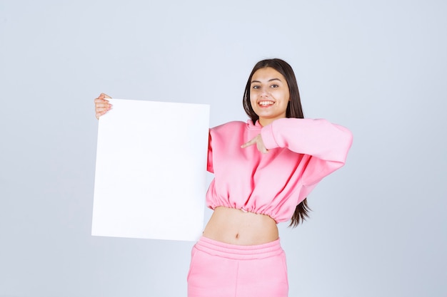 Foto grátis menina de pijama rosa, segurando uma placa de apresentação quadrada em branco e apontando para ela.