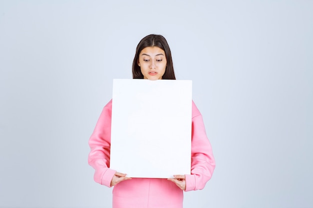 Menina de pijama rosa, segurando uma placa de apresentação quadrada em branco na frente dela.