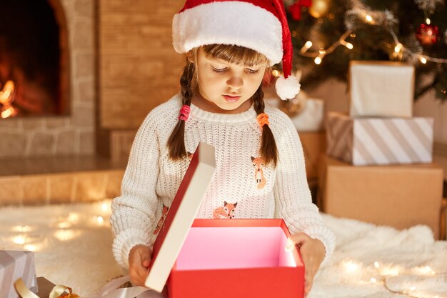 Menina de suéter branco e chapéu de Papai Noel, abrindo a caixa de presente com algo brilhando dentro, posando na sala festiva com lareira e árvore de Natal.