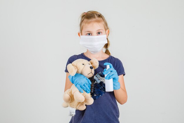 Foto grátis menina de t-shirt, luvas e máscara médica segurando spray de mão e urso, vista frontal.