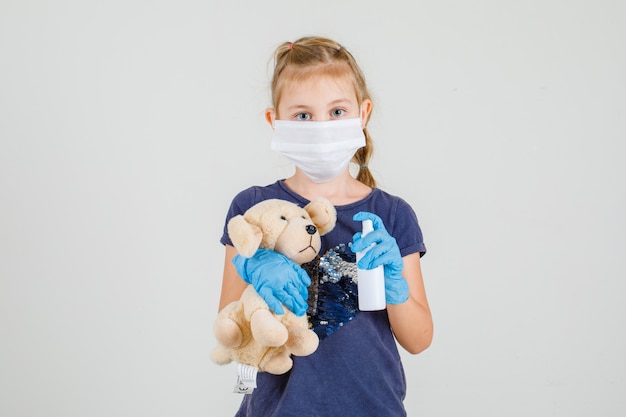 Menina de t-shirt, luvas e máscara médica segurando spray de mão e urso, vista frontal.