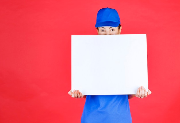 Menina de uniforme azul e boina, segurando uma mesa de informações quadrada branca e se sentindo positiva.