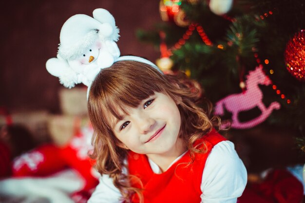 Menina de vestido vermelho senta com uma árvore de Natal