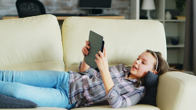 Menina deitada no sofá usando seu tablet. Criança alegre.