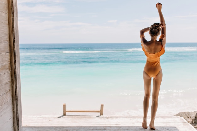 Menina descalça em maiô laranja, olhando para o oceano de manhã. Foto ao ar livre de modelo feminino relaxado com pele bronzeada, apreciando a vista para o mar.