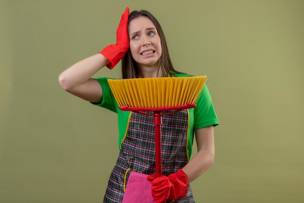 Menina descontente com a limpeza de uniforme com luvas vermelhas, segurando o esfregão e colocando a mão na cabeça na parede verde isolada