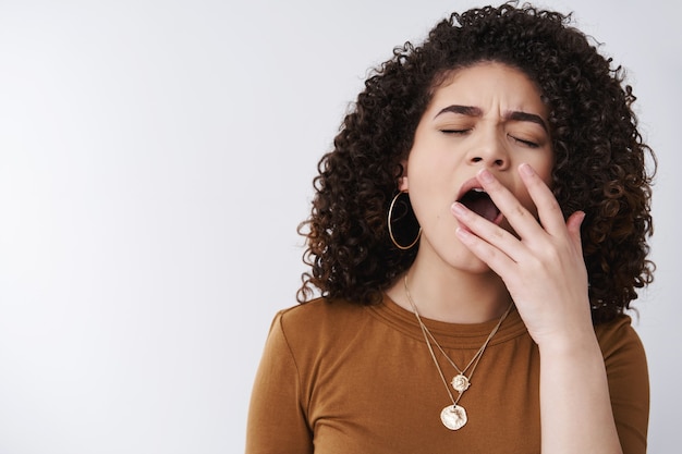 Menina desinteressada adormecer conversas chatas. Mulher bonita dos 20 anos de cabelo encaracolado bocejando olhos fechados, boca aberta, palma, sentindo sono, cansaço, tédio, preciso de café, levante-se de manhã cedo para trabalhar