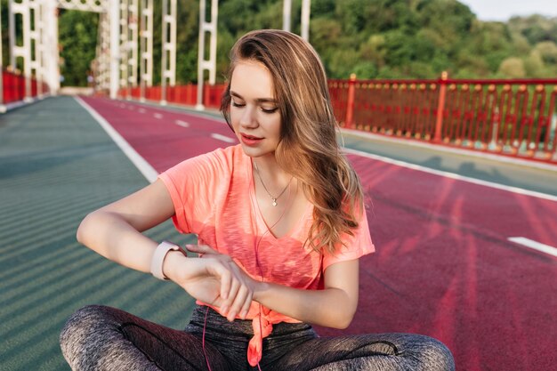 Menina desportiva sonhadora, olhando para o relógio após o treino. Tiro ao ar livre de mulher refinada relaxante antes da maratona no estádio.