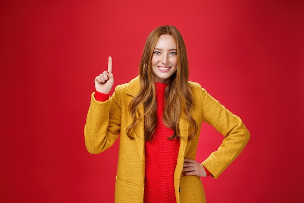 Menina dizendo a primeira razão, use o conselho dela. Mulher ruiva jovem energizada bonita e amigável no casaco amarelo, levantando a mão, mostrando o número um e sorrindo amplamente para a câmera contra o fundo vermelho.