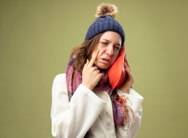 Menina doente chorando vestindo túnica branca e chapéu de inverno com lenço colocando uma bolsa de água quente na bochecha, segurando o dedo na bochecha isolado em verde oliva
