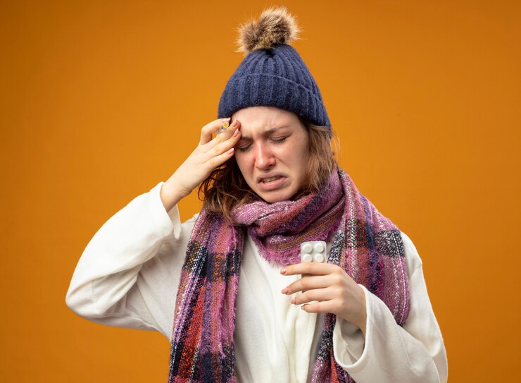 Foto grátis menina doente e dolorida vestindo túnica branca e chapéu de inverno com lenço segurando uma seringa com comprimidos colocando a mão na testa isolada na parede laranja