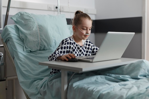 Menina doente jogando no laptop enquanto hospitalizada na enfermaria pediátrica da clínica de saúde. Criança doente descansando na cama do paciente enquanto desfruta de jogos no computador moderno.