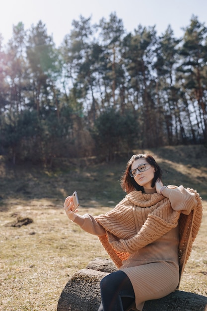 Menina elegante atraente jovem sobre a natureza na parede da floresta com um telefone em um dia ensolarado, tirando fotos de si mesma. férias ao ar livre e dependência de tecnologia.