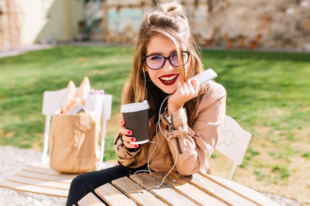 Menina elegante branca em copos grandes bebe café no parque e ouve música em fones de ouvido com interesse, olhando para a câmera. Pausa para o café no café ao ar livre, depois das compras.