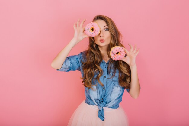 Menina elegante com cabelo longo cacheado posa positivamente, segurando rosquinhas rosa frescas com pó pronto para desfrutar de doces. Retrato de mulher jovem e atraente em camisa jeans retrô se divertindo com coisas doces