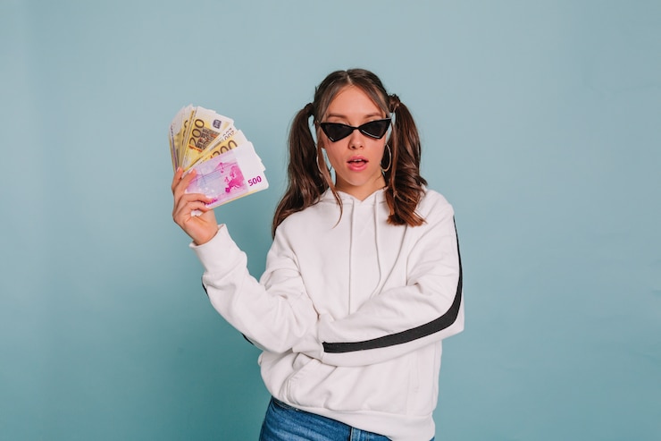 Foto grátis menina elegante com cabelos presos, usando óculos escuros pretos e pulôver branco, posando com um maço de dinheiro na parede isolada