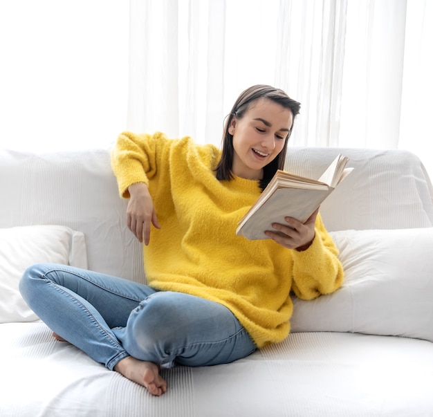 Menina elegante com um suéter amarelo está descansando em casa no sofá com um livro.