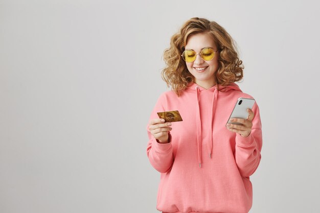 Menina elegante de cabelos crespos fazendo compras online, usando cartão de crédito e smartphone