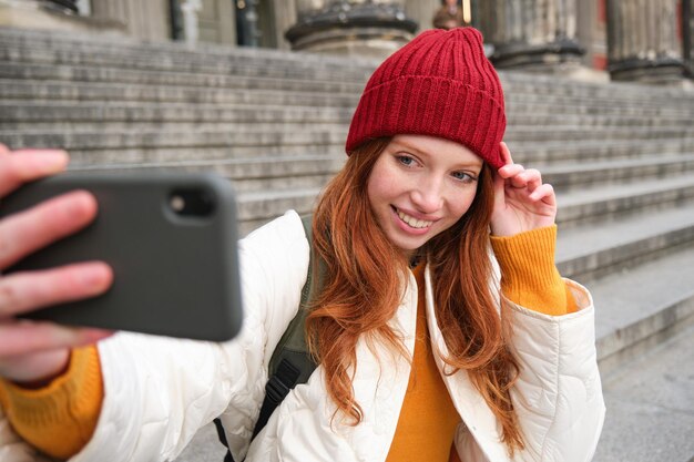Menina elegante de chapéu vermelho tira fotos na câmera do smartphone faz selfie enquanto ela se senta na escada n