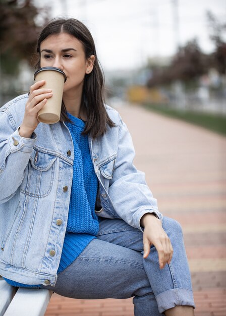 Menina elegante e alegre em estilo casual aprecia um café para viagem durante uma caminhada