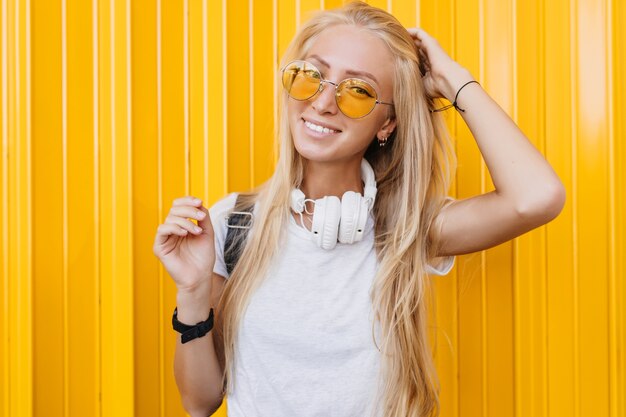 Menina elegante e graciosa brincando com seus longos cabelos loiros. espetacular jovem de óculos escuros e fones de ouvido.