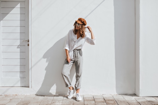 Menina elegante em calça cinza e blusa de algodão branca se passando perto de uma parede branca. mulher de boné e óculos.