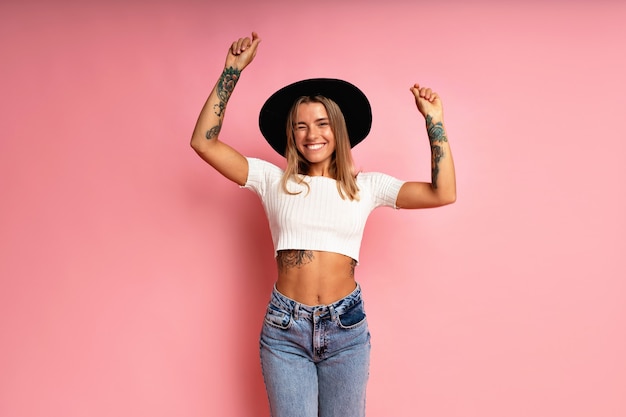 Foto grátis menina elegante hippie posando na parede rosa, com jeans crop top e fedora preto, humor alegre e brincalhão.