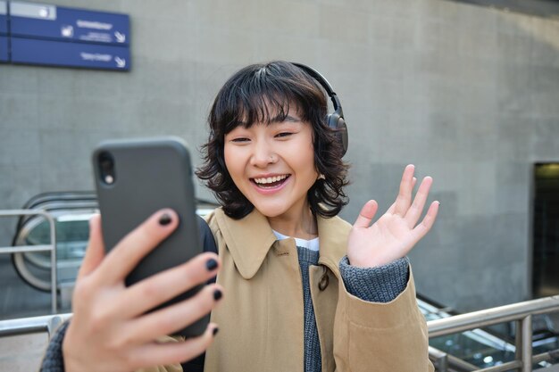 Menina elegante hipster em fones de ouvido diz olá para a câmera do celular acena com a mão no vídeo do smartphone