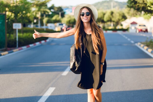 Menina elegante pedindo carona na estrada usando vestido preto curto, chapéu de palha, óculos pretos e mochila preta. Ela sorri sob os raios quentes do sol poente