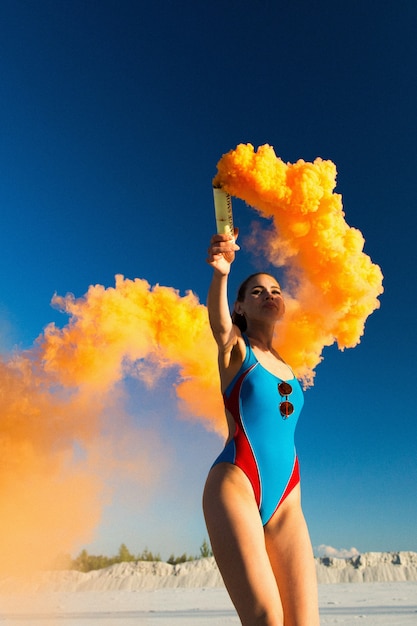 Menina em danças azuis de nadar-traje com fumaça de laranja na praia branca