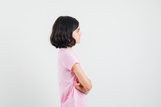 Menina em pé com os braços cruzados em uma camiseta rosa e olhando com foco.