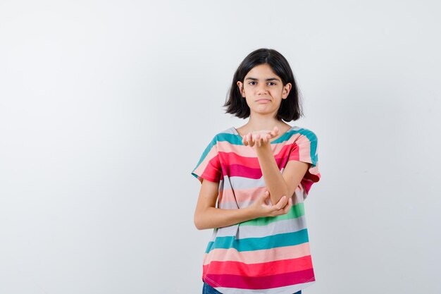 Menina em t-shirt, esticando a mão como segurando algo, mantendo uma mão no cotovelo e parecendo descontente, vista frontal.