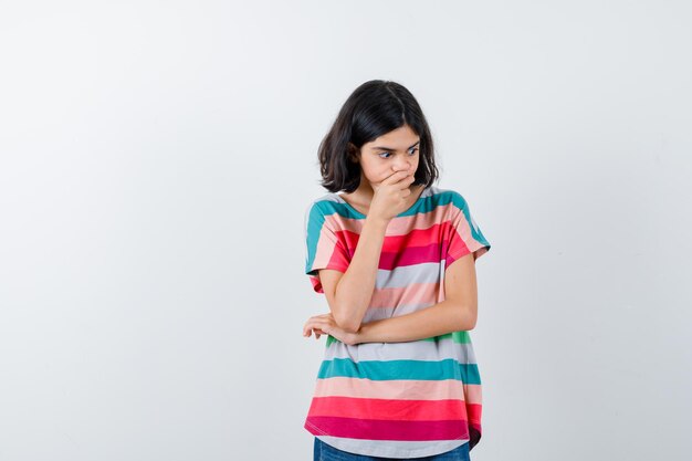 Menina em t-shirt, mantendo a mão no queixo e olhando pensativa, vista frontal.