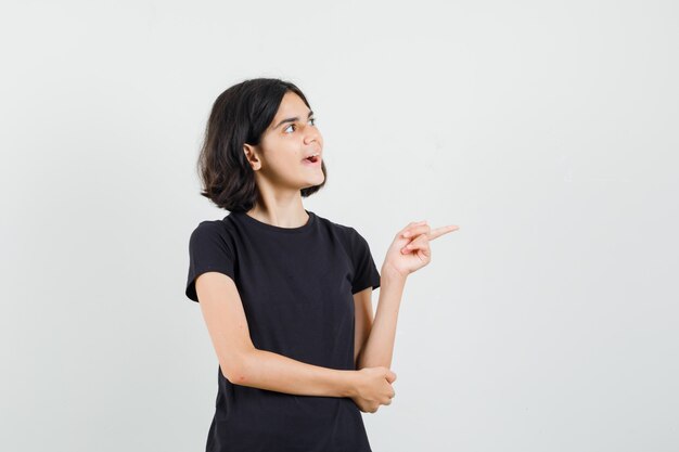 Menina em t-shirt preta apontando para o lado e olhando alegre, vista frontal.