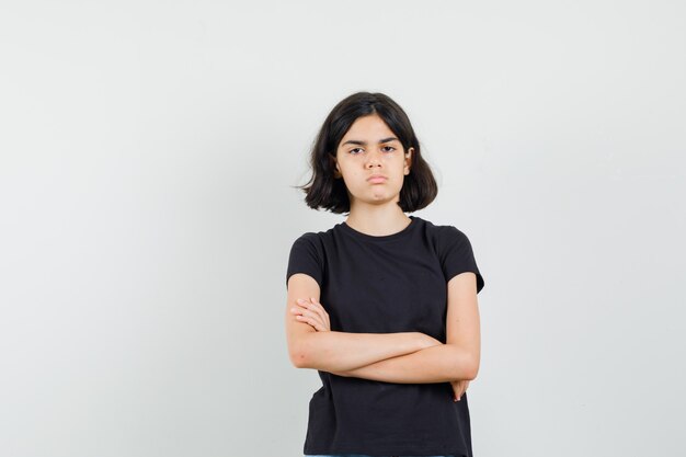 Menina em t-shirt preta em pé com os braços cruzados e olhando séria, vista frontal.