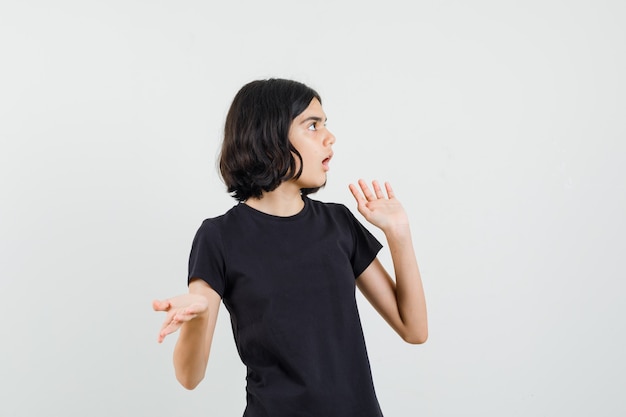 Menina em t-shirt preta, mantendo as mãos em um gesto perplexo, vista frontal.