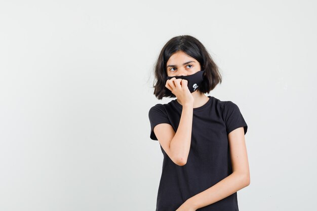 Menina em t-shirt preta, máscara em pé em pose de pensamento e olhando sensata, vista frontal.