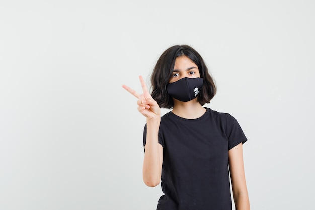 Menina em t-shirt preta, máscara mostrando o sinal V e olhando confiante, vista frontal.