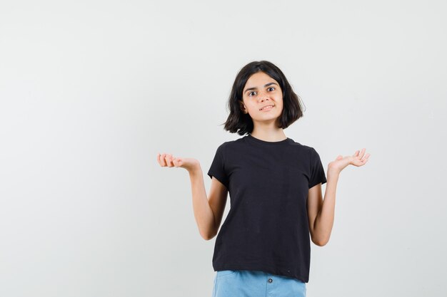 Menina em uma camiseta preta, shorts mostrando um gesto de impotência e parecendo confusa, vista frontal.