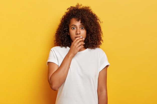 Menina emocional assustada de pele escura com cabelos crespos luminosos, cobre o rosto e olha com olhos esbugalhados, olha com expressão apavorada, temerosa pela própria vida, prende a respiração de excitação