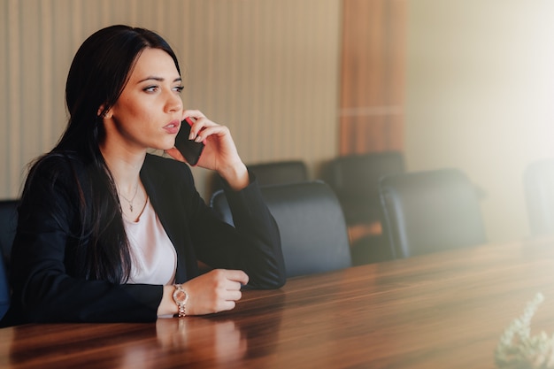 Foto grátis menina emocional atrativa nova na roupa do estilo do negócio que senta-se na mesa com o telefone no escritório ou na audiência